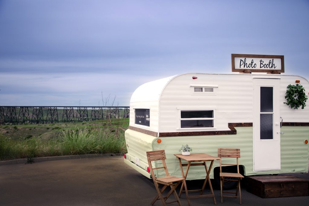 Vintage photo booth trailer overlooking a valley, with a wooden table and chairs placed beside it on a paved surface.