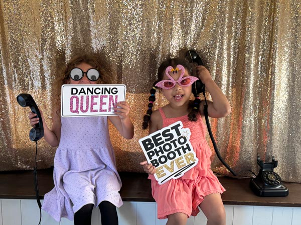 Two young girls dressed in cute dresses pose in a photo booth with a glittery gold backdrop. They are holding fun props, including a sign that says 'Dancing Queen' and another that reads 'Best Photo Booth Ever.' Both girls are wearing playful glasses, adding to the whimsical atmosphere.