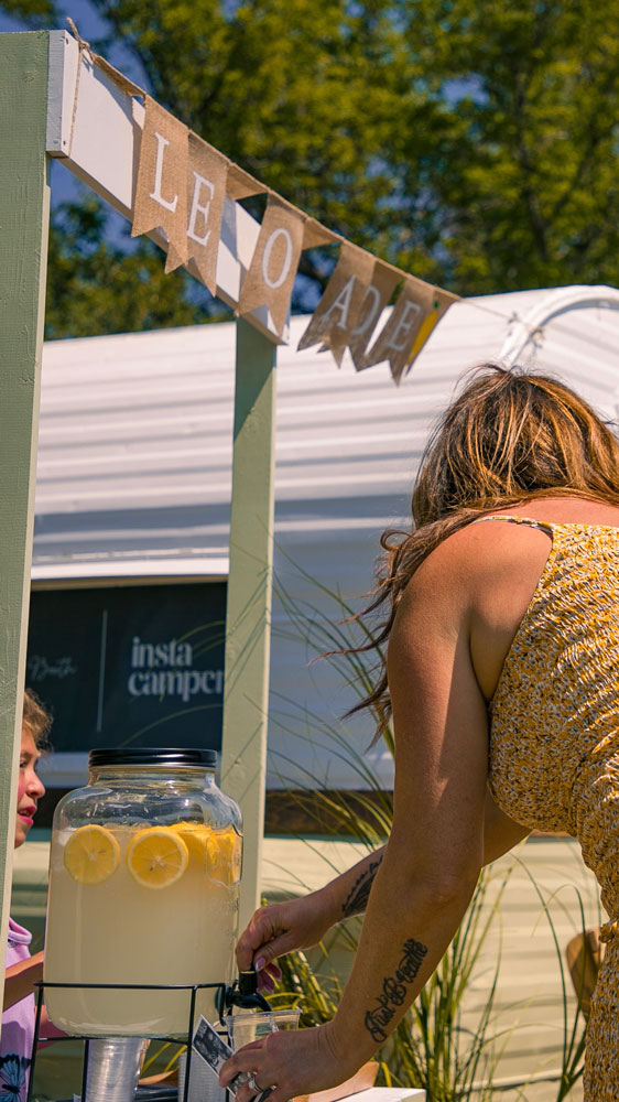 Woman pouring lemonade from a large glass dispenser at an outdoor lemonade stand, with the Instacamper trailer visible in the background.