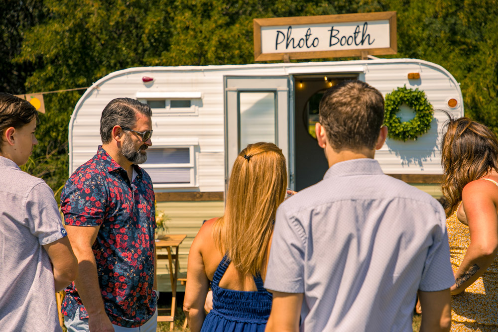 Group of people standing outside a vintage photo booth trailer at an outdoor event on a sunny day.