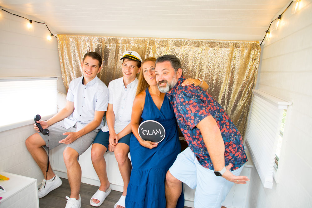 Family of four posing together inside a photo booth trailer with a gold sequin backdrop, holding props including a “GLAM” sign and a captain’s hat.